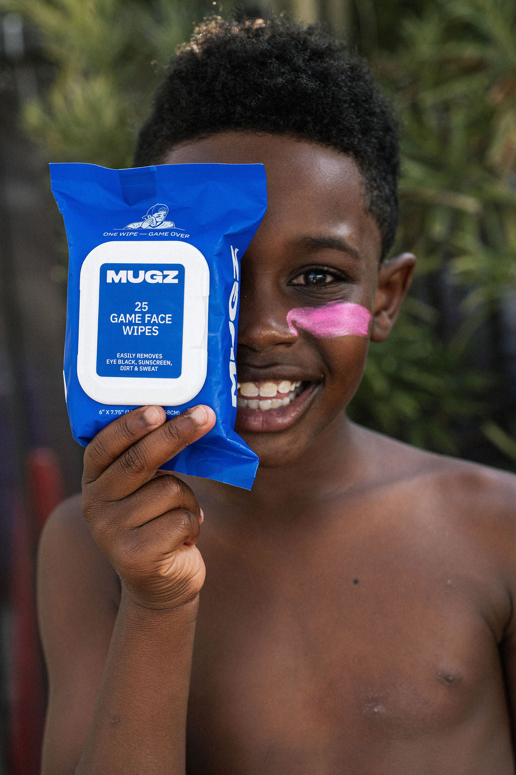 A smiling young boy with a pink zinc sunscreen holds up a blue package of MUGZ Game Face Wipes in front of his face.