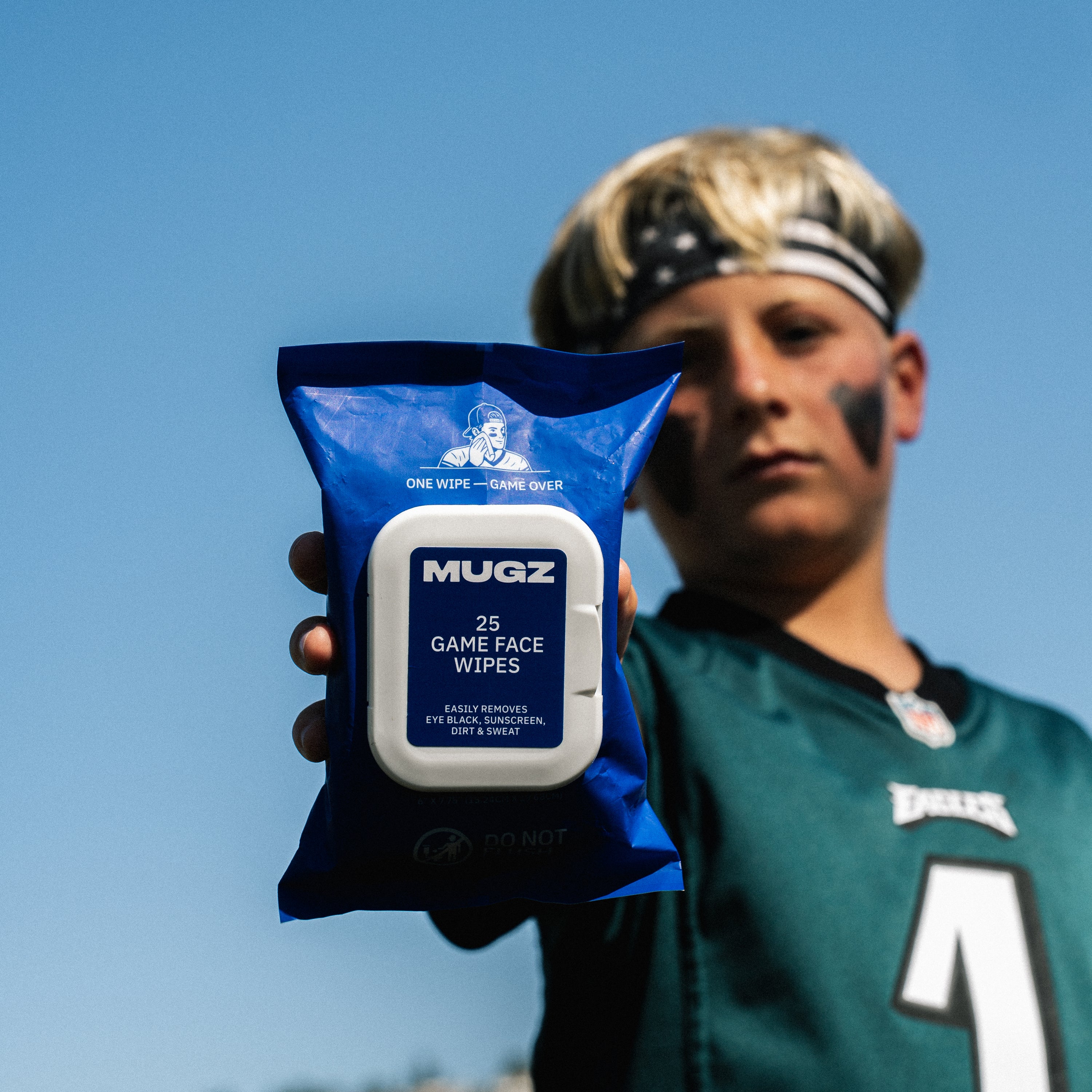 A football player in a green football jersey holds up a package of MUGZ Game Face Wipes against a clear blue sky background.